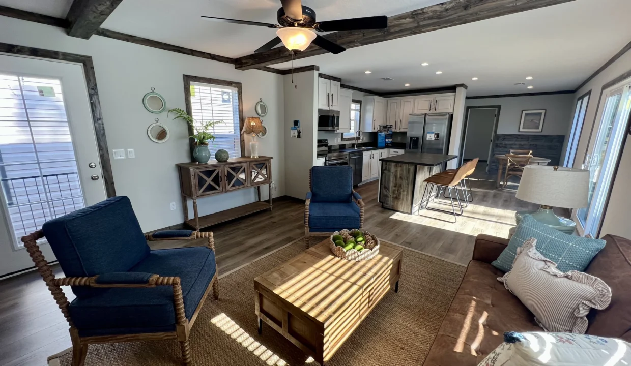 Modern living room interior of a Bandit manufactured home, featuring blue chairs, wooden coffee table, kitchen area with stainless steel appliances, and natural light streaming through windows.