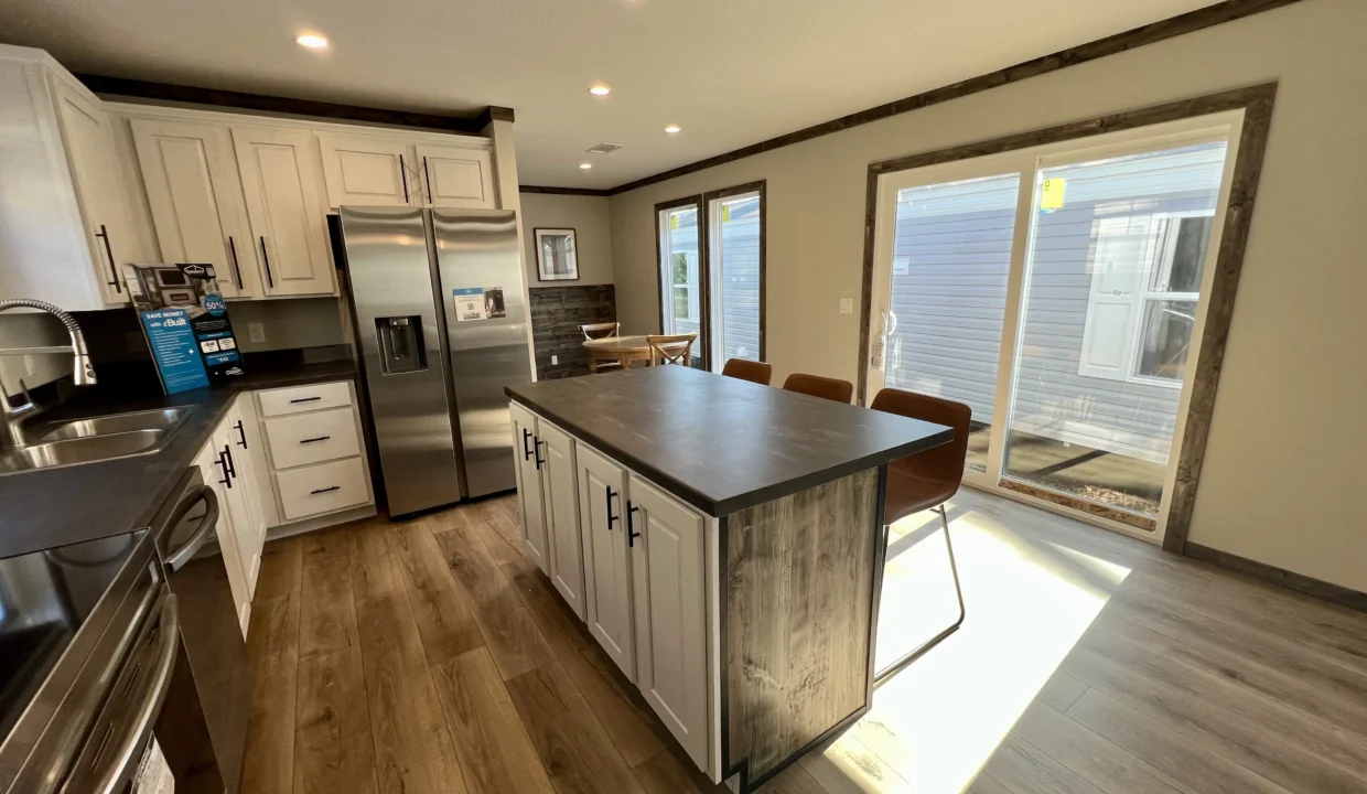 Modern kitchen interior featuring a central island, stainless steel refrigerator, white cabinetry, and natural light from sliding glass doors, highlighting the Bandit manufactured home by Sanders Housing.