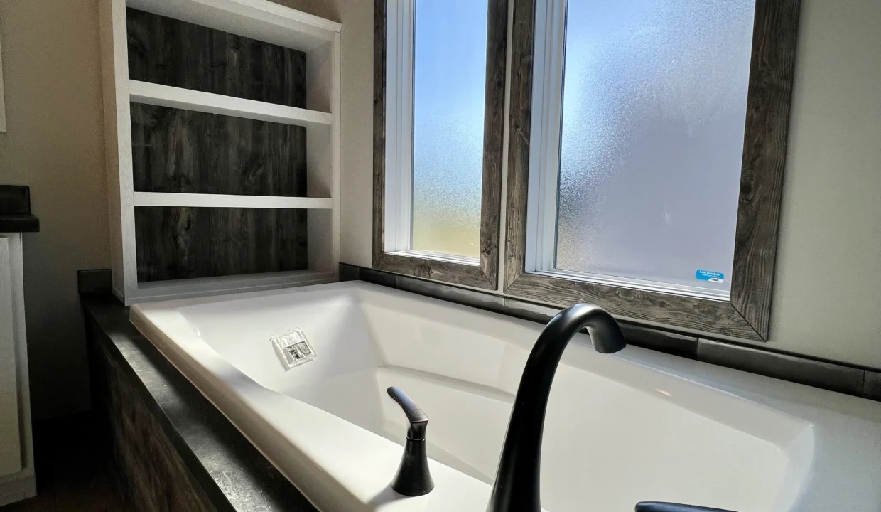 Modern bathtub with black faucet and wooden shelving, featuring large windows for natural light, highlighting the Bandit manufactured home&rsquo;s bathroom design.