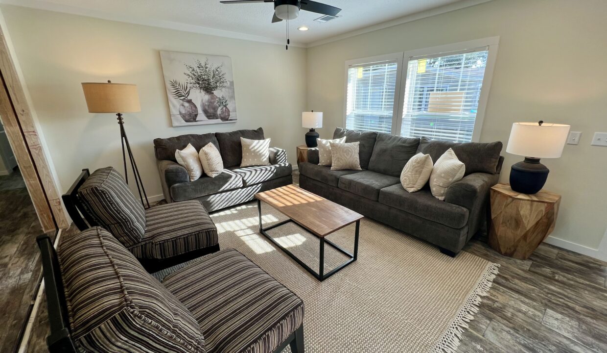 Living room interior of the Boujee XL manufactured home featuring a cozy seating arrangement with dark gray sofas, decorative pillows, a wooden coffee table, and modern lighting.
