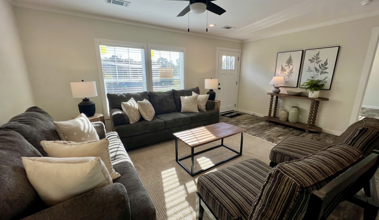 Living room interior featuring a spacious layout with a dark gray sofa adorned with decorative pillows, a wooden coffee table, and stylish armchairs, complemented by natural light from large windows and modern decor elements.
