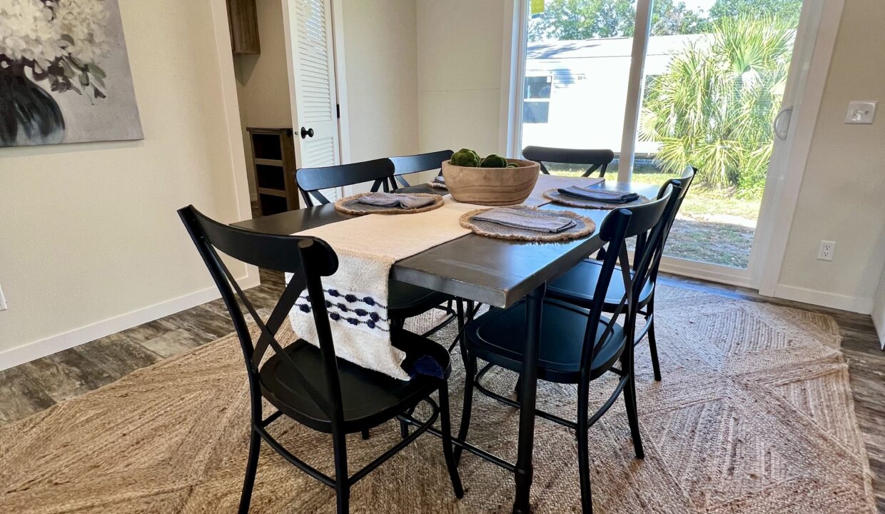 Dining area in Boujee XL manufactured home featuring a black table with decorative placemats, a bowl of green fruit, and a view of outdoor greenery through large windows.