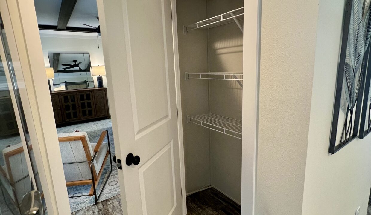 Interior view of a closet with white wire shelving, showcasing a spacious design, with a glimpse of a living area featuring a wooden cabinet and lamps in the background, relevant to the Boujee XL manufactured home by Sanders Housing.