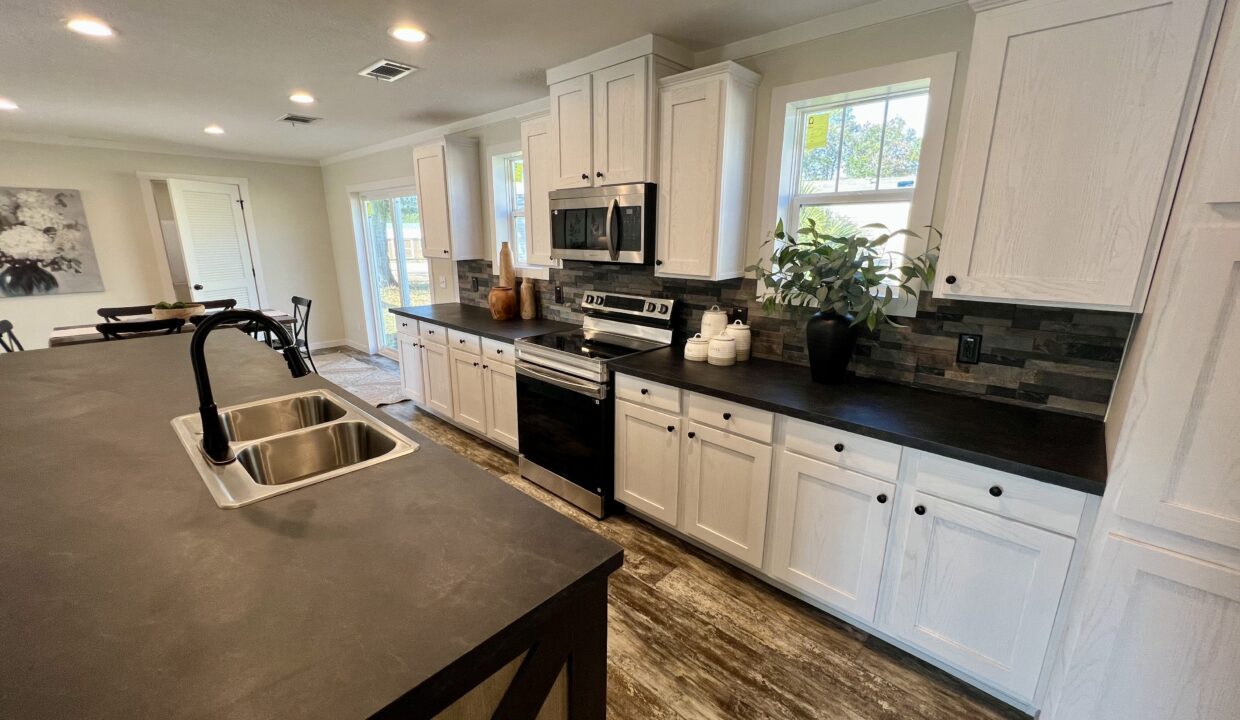 Modern kitchen in Boujee XL manufactured home featuring white cabinetry, stainless steel appliances, and a dark countertop, showcasing spacious design and gourmet features.