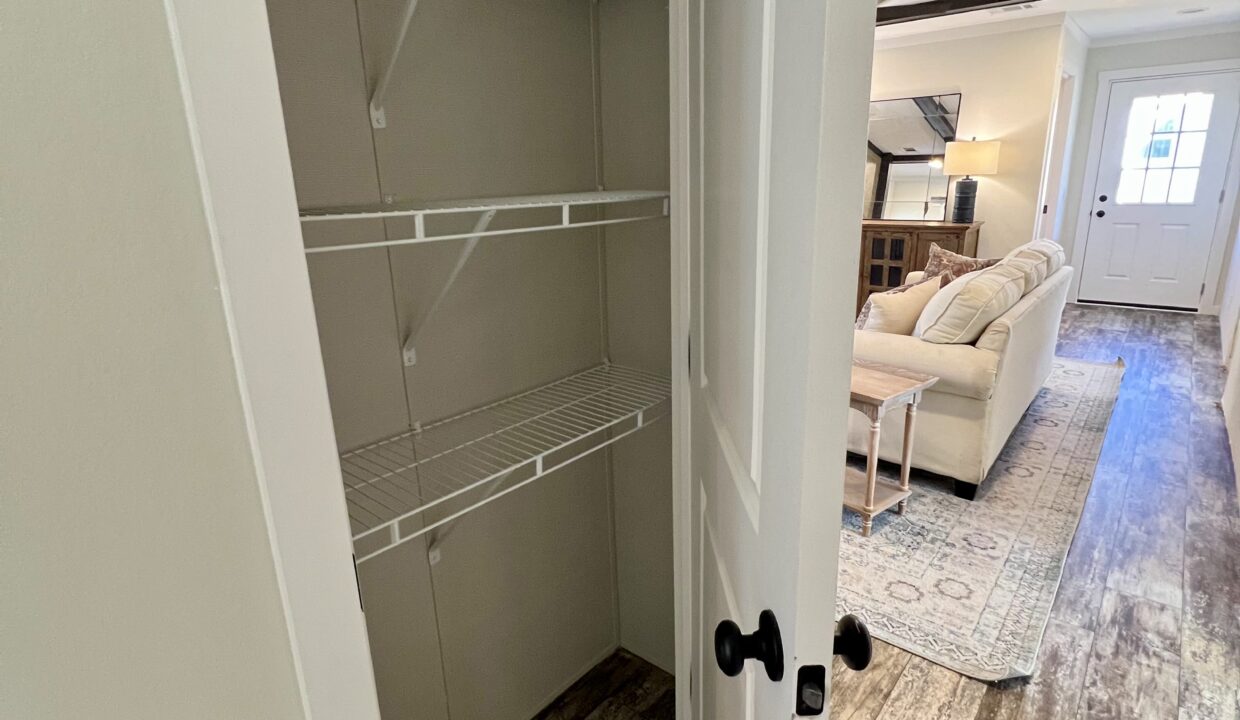 Interior view of a storage closet with wire shelving, showcasing a cozy living area with a sofa and coffee table in the background, highlighting the spacious design of the Boujee XL manufactured home by Sanders Housing.