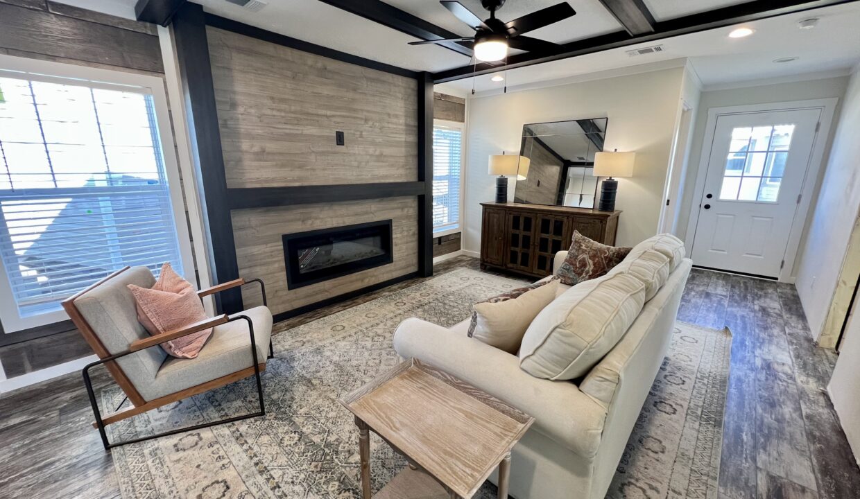 Modern living room interior of Boujee XL manufactured home, featuring a cozy sofa, stylish armchair, decorative pillows, wall-mounted television, electric fireplace, and natural light from large windows.