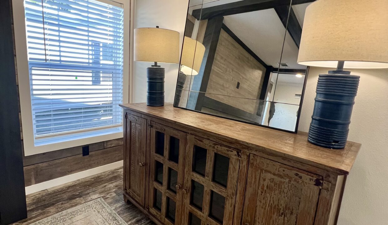 Modern interior of a manufactured home featuring a wooden cabinet, decorative mirror, and stylish lamp beside a window with blinds.