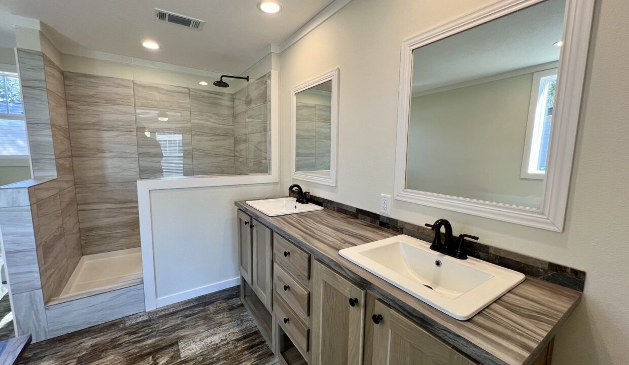 Luxurious bathroom interior featuring dual sinks, modern fixtures, spacious shower area with tile walls, and ample natural light from windows, showcasing the Boujee XL manufactured home by Sanders Housing.