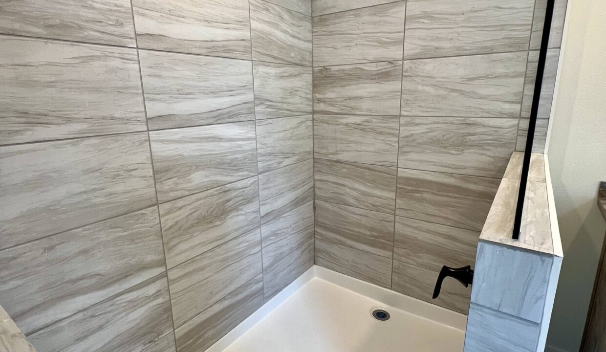 Modern shower featuring elegant marble-like tiles and a sleek black faucet, showcasing the luxurious design of the Boujee XL manufactured home by Sanders Housing.