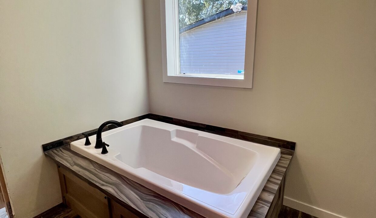 Luxurious bathroom featuring a spacious white bathtub with modern black faucet, natural light from a window, and elegant countertop design, highlighting the Boujee XL manufactured home by Sanders Housing.