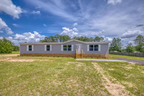 Multi-section home for sale by Sanders Housing, featuring a light gray exterior, large windows, and a welcoming front porch, set against a blue sky with scattered clouds.