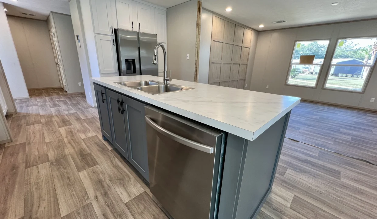 Modern kitchen interior featuring a spacious island with a double sink, stainless steel dishwasher, and light wood flooring, showcasing the Snowcap manufactured home by Sanders Housing.