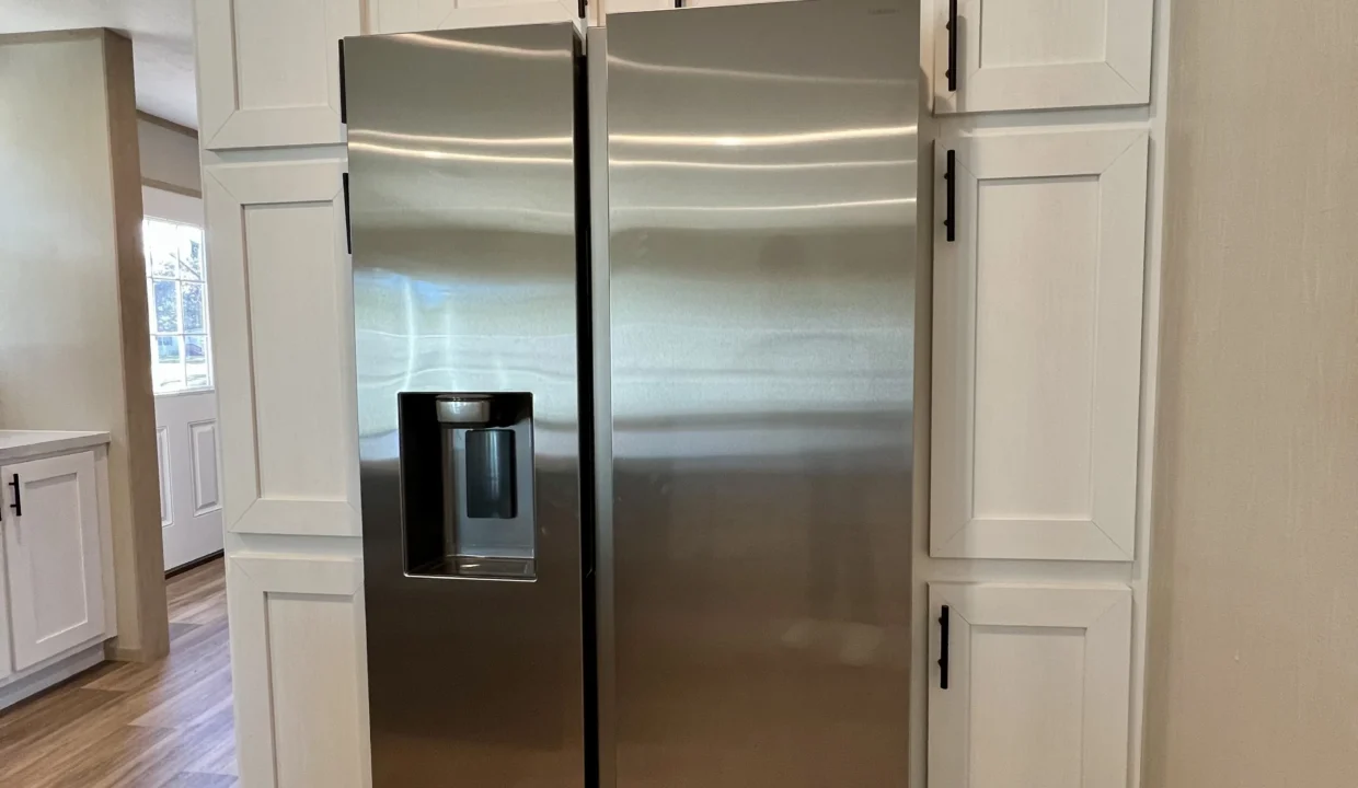Stainless steel refrigerator with water dispenser, adjacent to white cabinetry in modern kitchen setting, showcasing amenities of the Snowcap manufactured home by Sanders Housing.