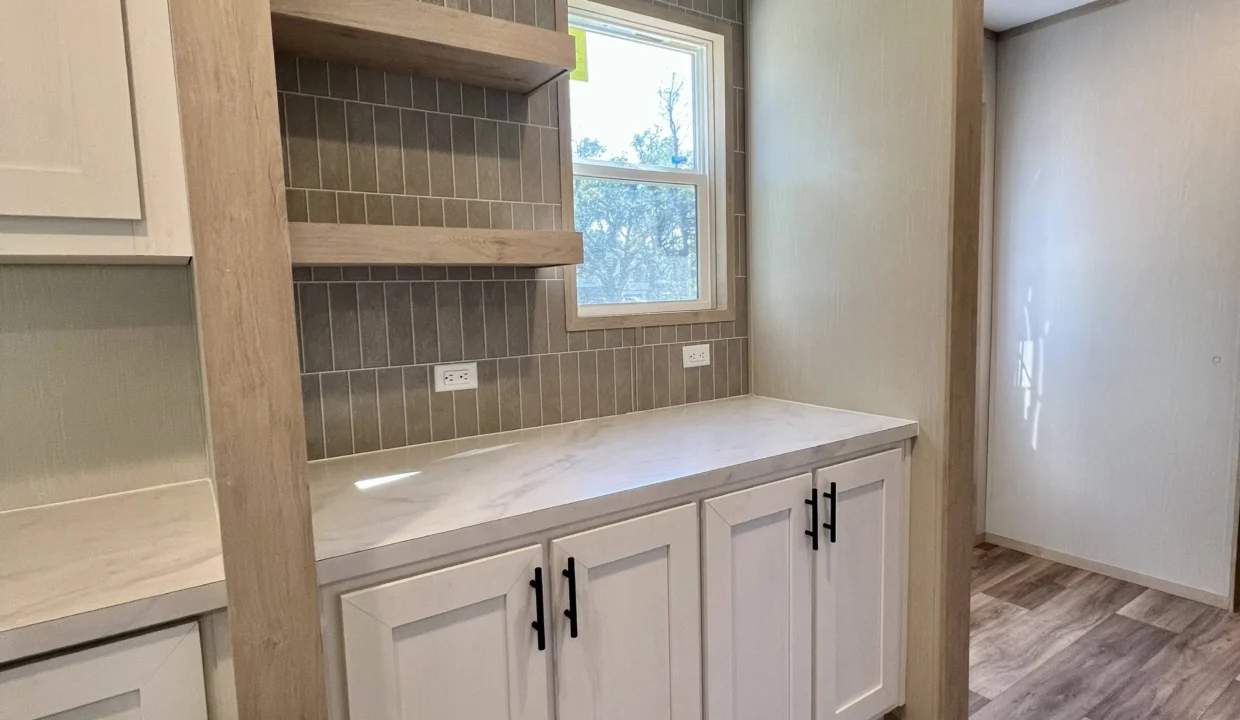 Modern kitchen interior featuring white cabinetry, open shelving, and a marble countertop, showcasing the spacious design and contemporary style of the Snowcap manufactured home by Sanders Housing.