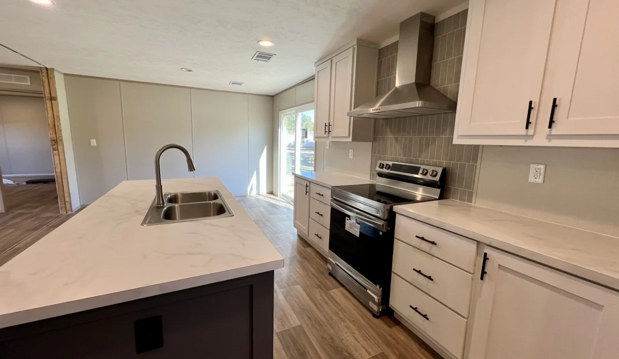Modern kitchen interior featuring a spacious island with a sink, stainless steel appliances, white cabinetry, and natural light from large windows, showcasing the Snowcap manufactured home by Sanders Housing.