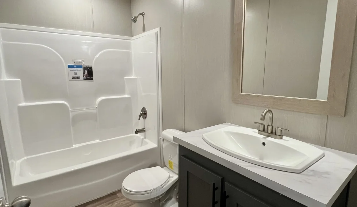 Modern bathroom featuring a white bathtub and shower combination, sleek sink with faucet, and a large mirror, showcasing the interior design of the Snowcap manufactured home by Sanders Housing.