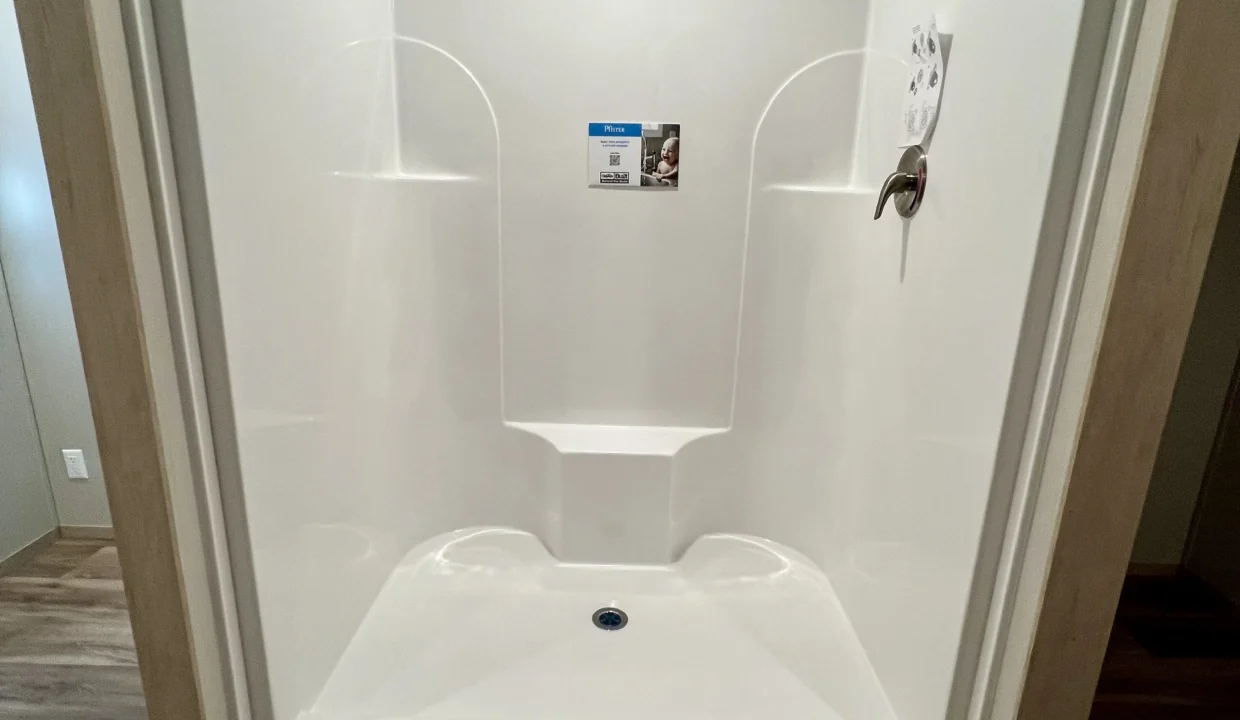 White shower stall with built-in seating, featuring a product label, showcasing modern amenities in the Snowcap manufactured home by Sanders Housing.