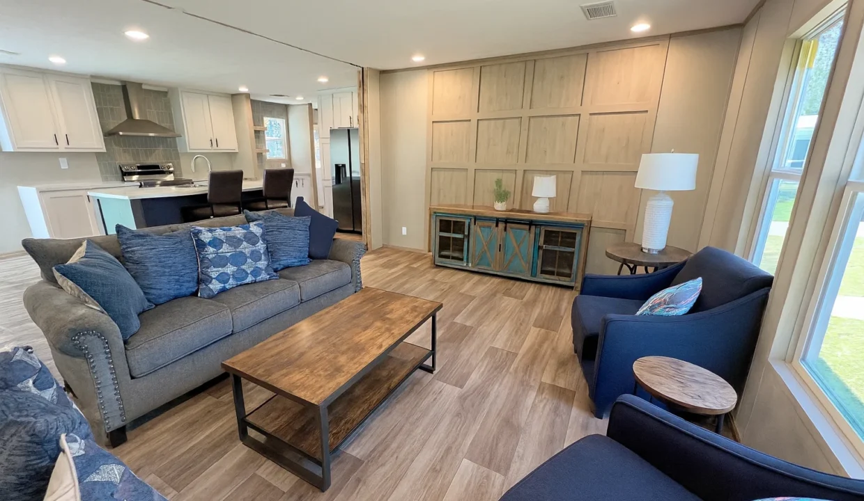 Spacious living room of the Snowcap manufactured home featuring a gray sofa with decorative pillows, wooden coffee table, stylish blue chairs, and modern kitchen area in the background.