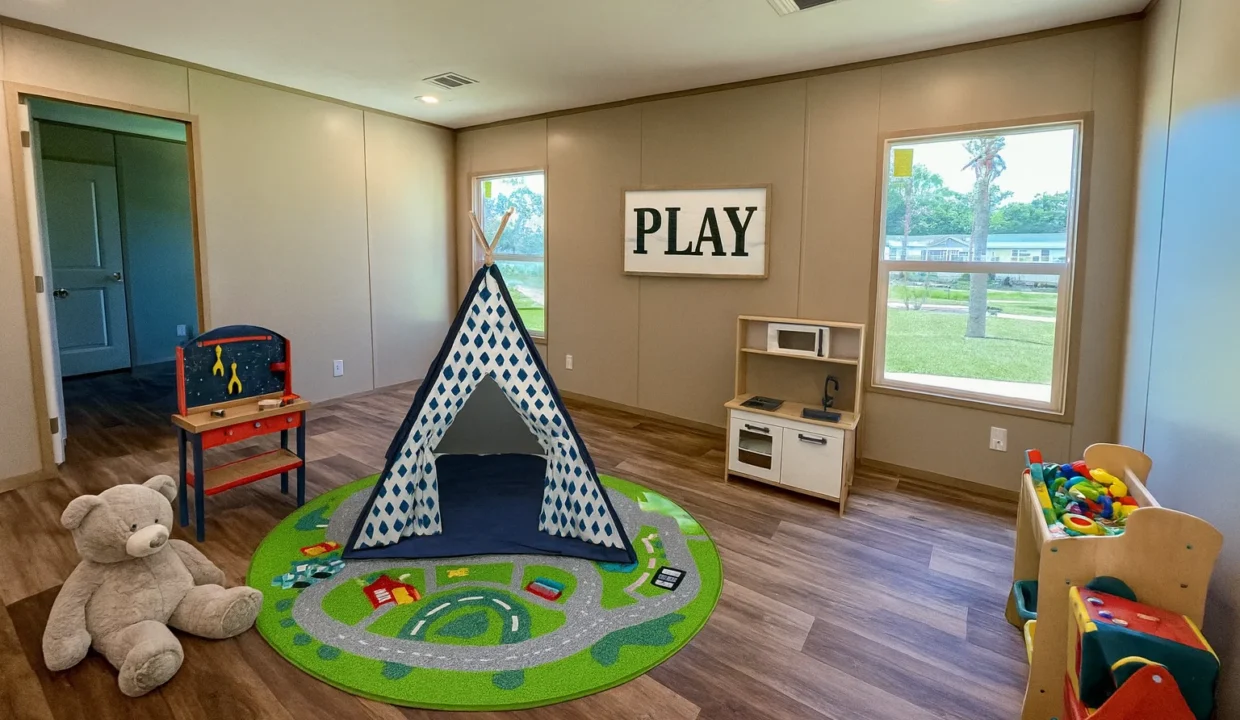Playroom featuring a teepee, colorful rug with road design, plush teddy bear, play kitchen, and a sign reading "PLAY," showcasing a family-friendly space in the Snowcap manufactured home by Sanders Housing.